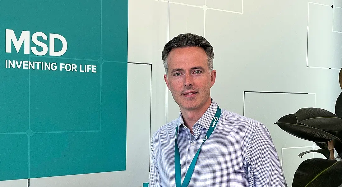 A man in a light blue shirt and a green lanyard smiles at the camera. Beside him is a sign with the MSD logo.