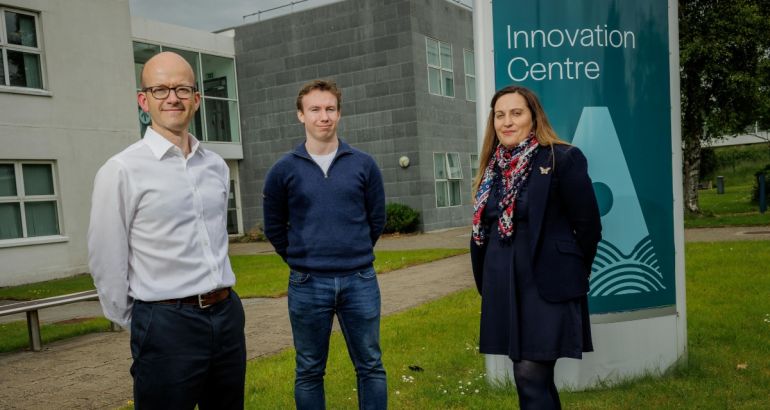 Two men and a woman standing outside a grey building with grass under them and a grey sky overhead. There is a sign behind the woman that reads "Innovation Centre".