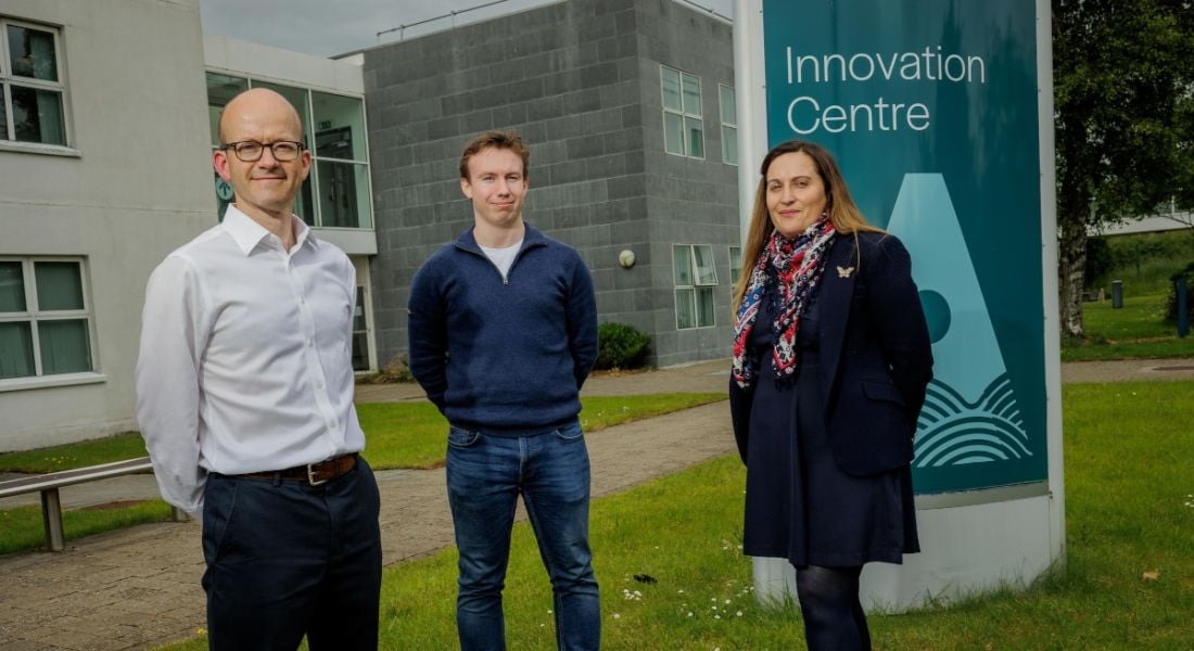 Two men and a woman standing outside a grey building with grass under them and a grey sky overhead. There is a sign behind the woman that reads "Innovation Centre".