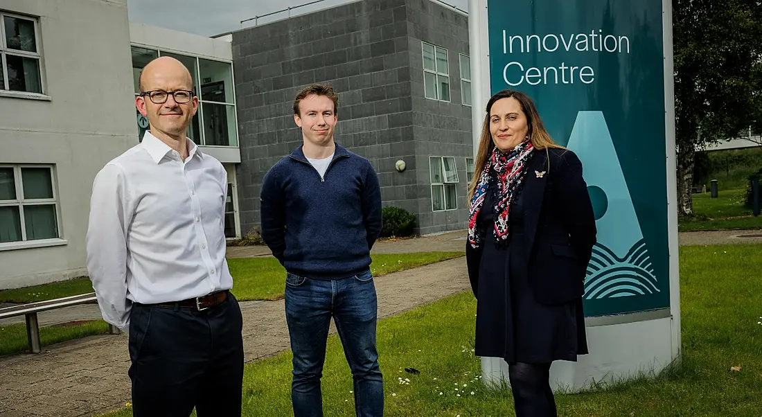 Two men and a woman standing outside a grey building with grass under them and a grey sky overhead. There is a sign behind the woman that reads "Innovation Centre".