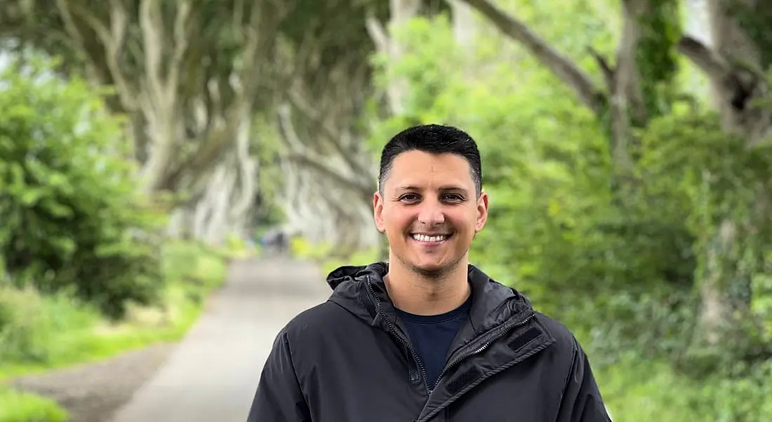 A young man wearing a black rain jacket stands in the middle of a path with trees and bushes either side. He is smiling at the camera.