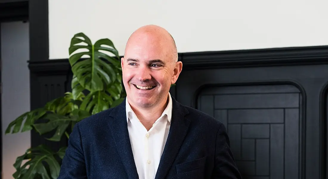 Darragh McCarthy looking away from the camera and smiling with an indoor plant in the background.