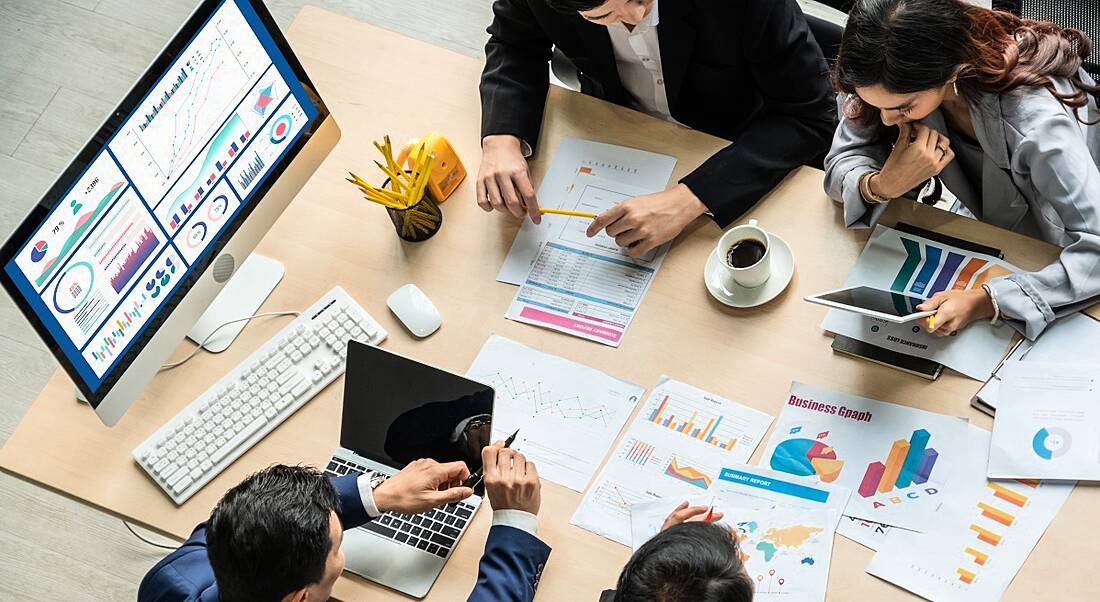 An aerial view of four people sitting around a table with screens and pieces of paper showing various types of data analytics.