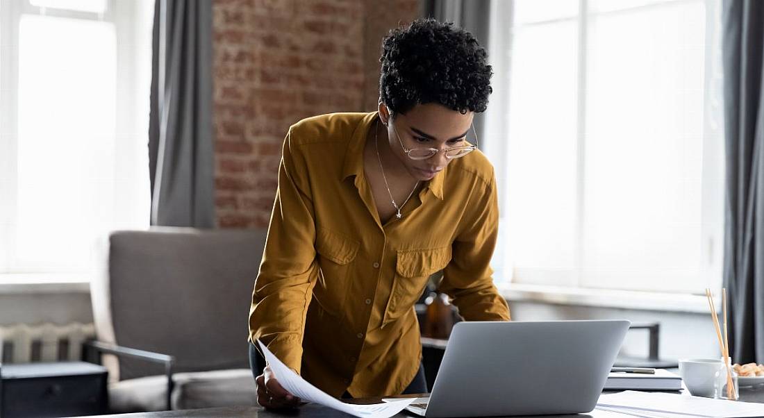 A woman standing at a desk working at a laptop in a brightly lit room. She’s wearing a yellow shirt and glasses.