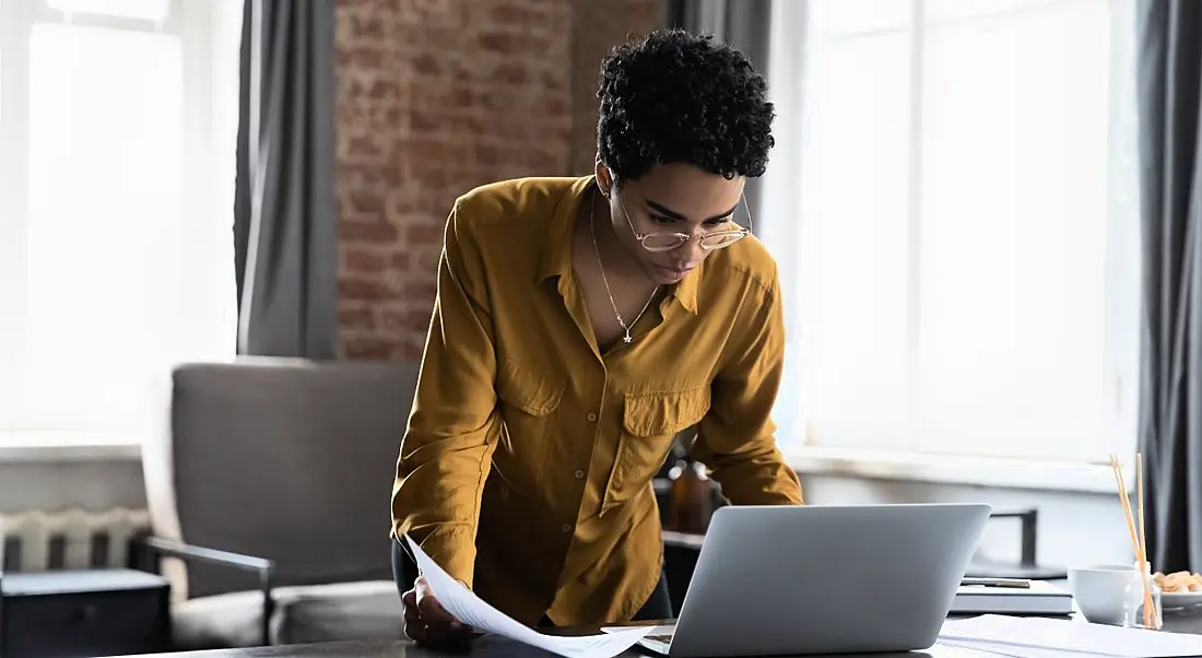 A woman standing at a desk working at a laptop in a brightly lit room. She’s wearing a yellow shirt and glasses.