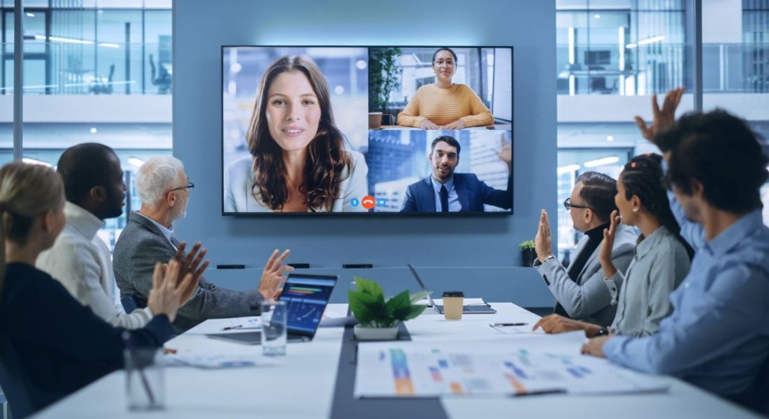 A boardroom meeting with a large screen showing three remote employees on a conference call, symbolising hybrid meetings.