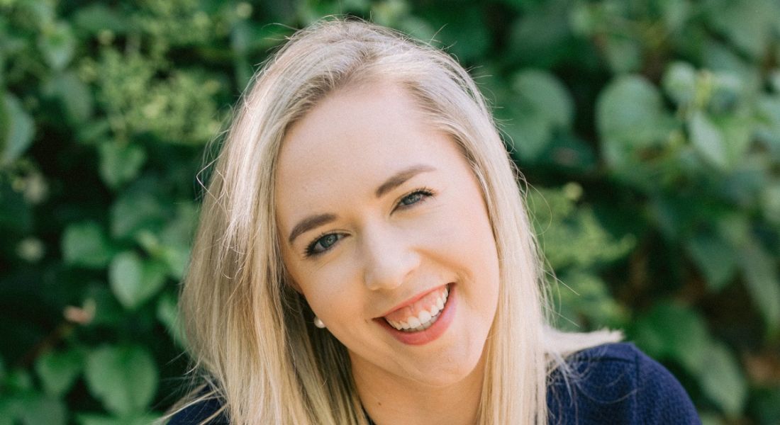 A smiling woman with blonde hair against a backdrop of green leaves. She is Andrea Graham, senior software engineering director at Workvivo.