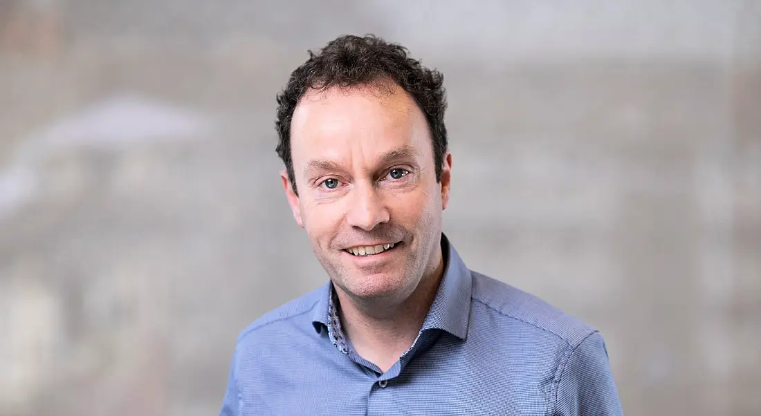 A headshot of a man with dark hair smiling at the camera. He is wearing a blue shirt against a grey background.