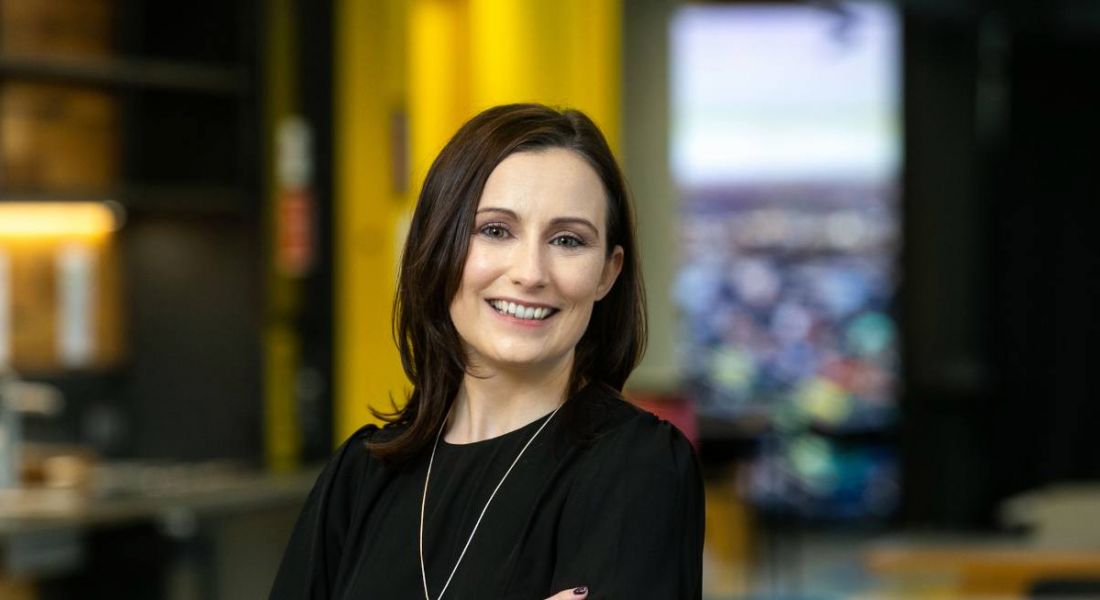 A woman with dark hair wearing a black dress smiles at the camera against the backdrop of a modern Accenture office.