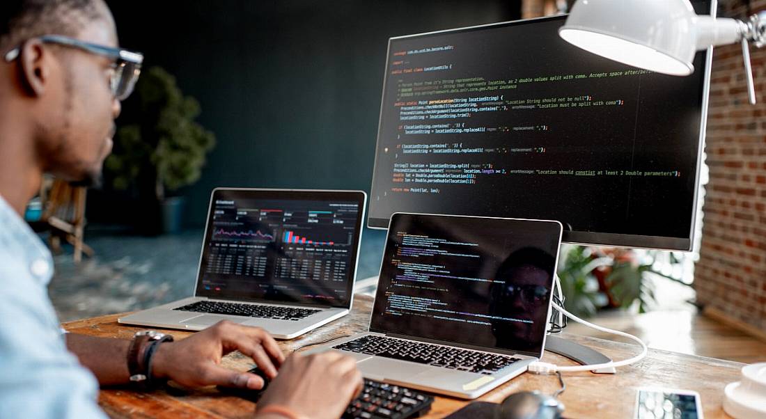 Man sitting at a desk with two laptops and a large screen monitor working as a software developer.