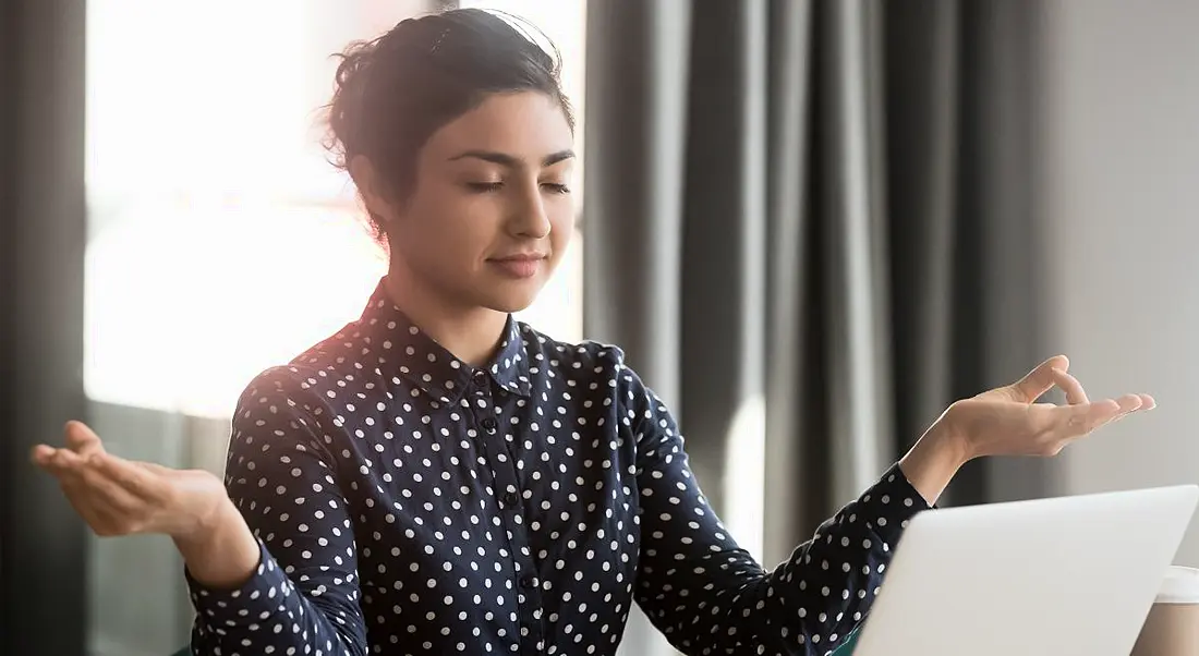 Woman making serene gesture with her hands while she works at a table with a laptop.