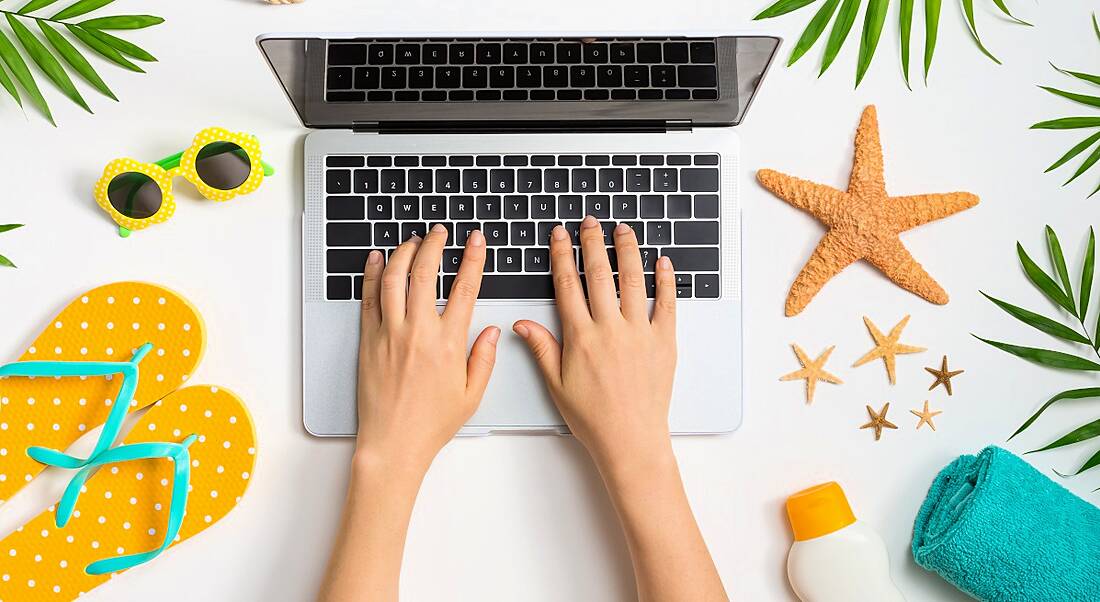 Person's hands working on a laptop with summer items like sandals and suncream and sunglasses surrounding it.