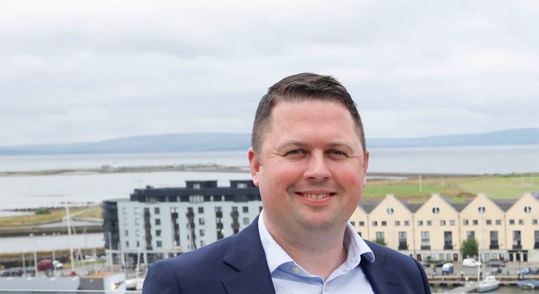 A headshot of Ruairi Conroy in front of the docks with buildings and the Galway skyline behind him.