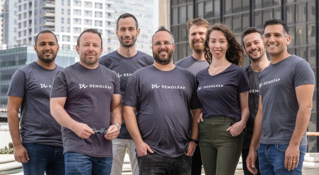 A group of people wearing grey T-shirts saying Demoleap on them standing in front of some buildings in a built-up urban area.