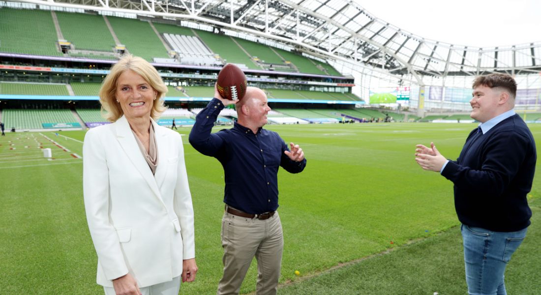 A woman and two men standing in the middle of a stadium. The woman is looking at the camera while the two men are about to pass an American football to each other.