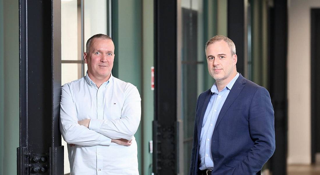 Two men in business clothing standing together in an office hallway, with windows and doors in the background.