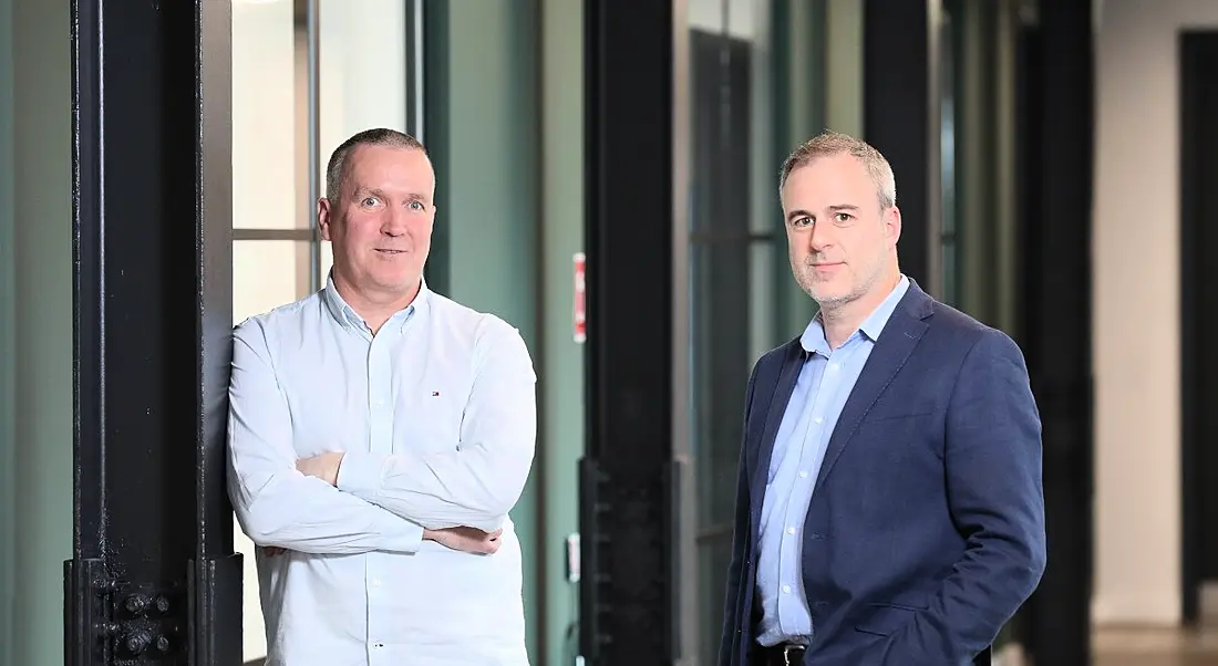 Two men in business clothing standing together in an office hallway, with windows and doors in the background.