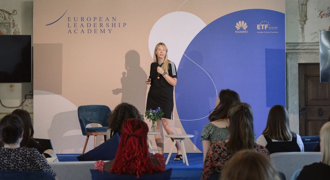 A woman wearing a black dress standing on a stage speaking to a room full of women. Behind her is a sign that says European Leadership Academy.