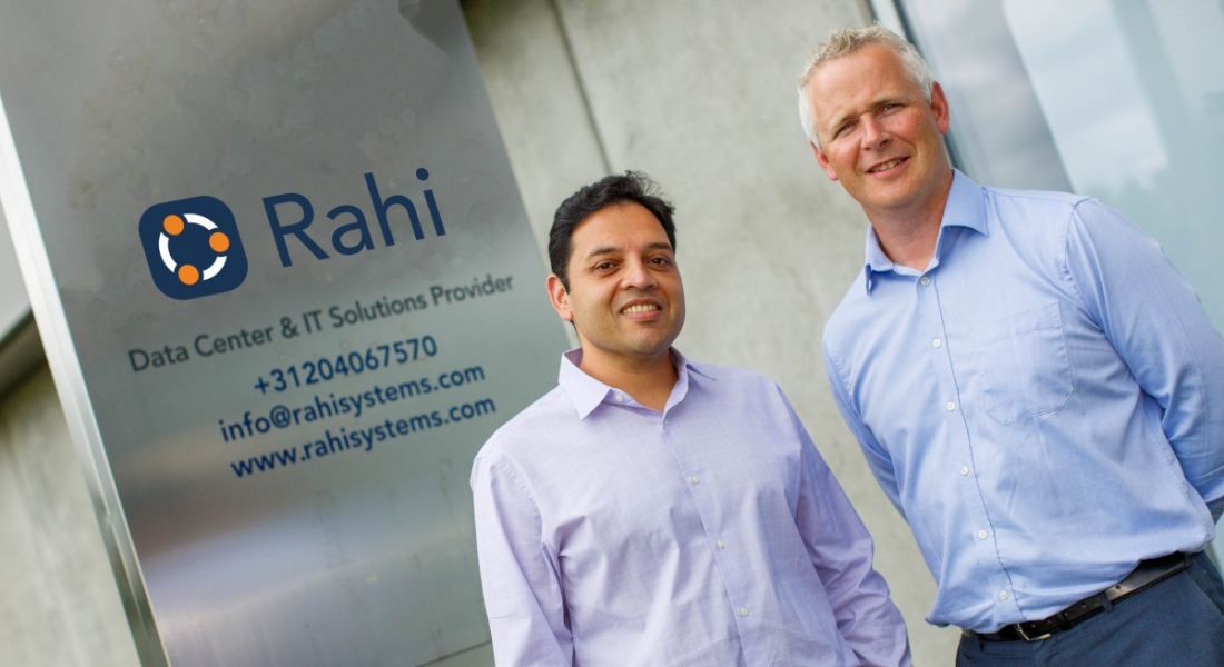 Two men standing in front of a silver sign for Rahi.