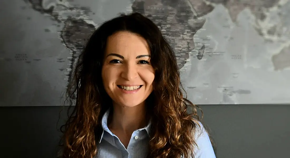 A young woman with dark hair smiles at the camera against a grey wall.