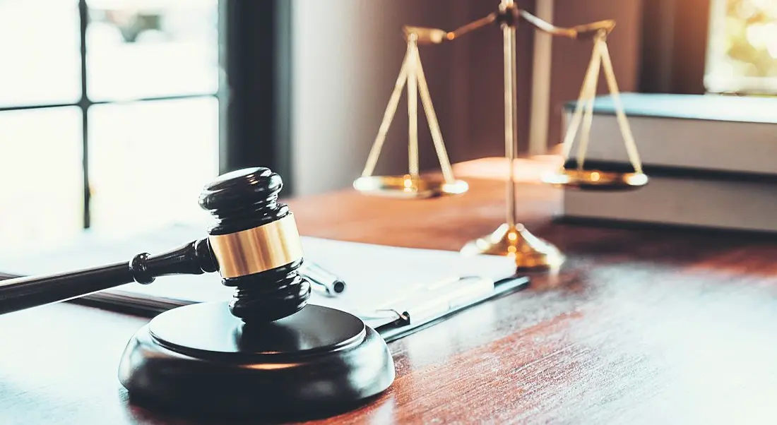 A judge’s gavel resting on the table. IN the background is the scales of justice, symbolising legal issues.