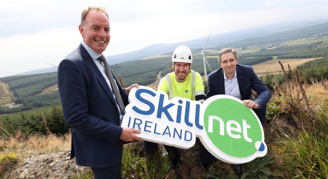 Three men holding a sign for Skillnet Ireland. They are all outdoors in a boggy area.