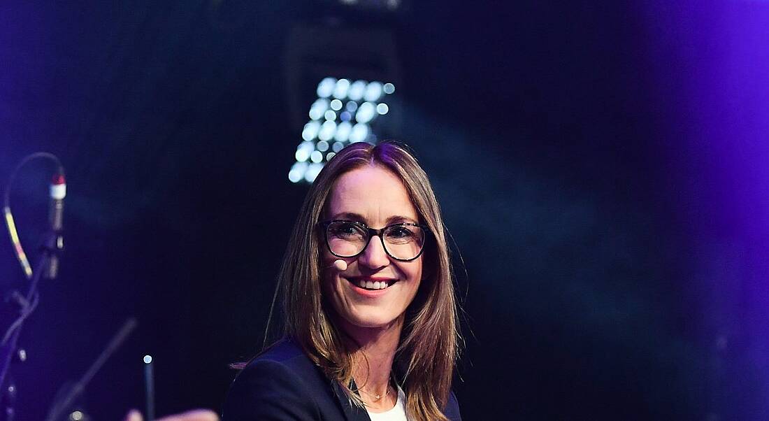 A young woman wearing a microphone smiling at someone off camera at an event.