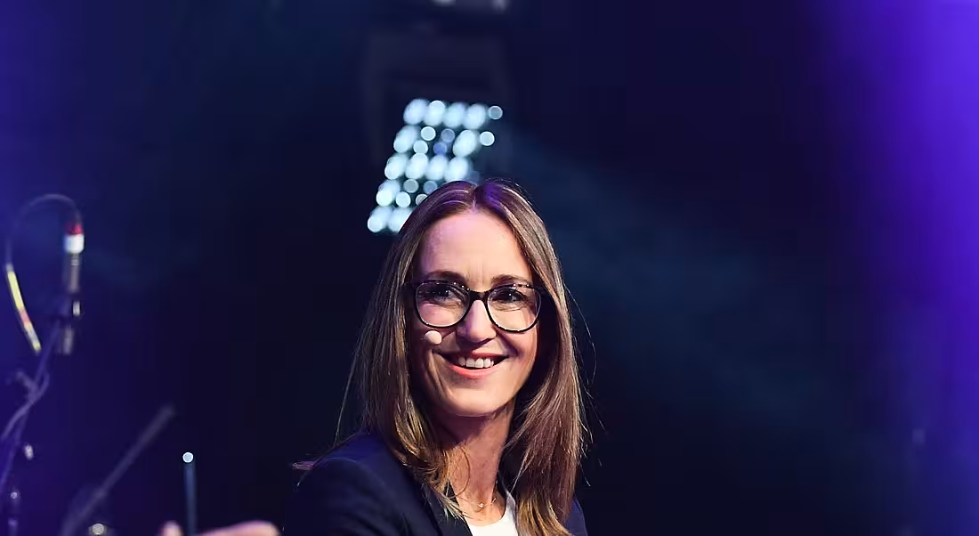 A young woman wearing a microphone smiling at someone off camera at an event.