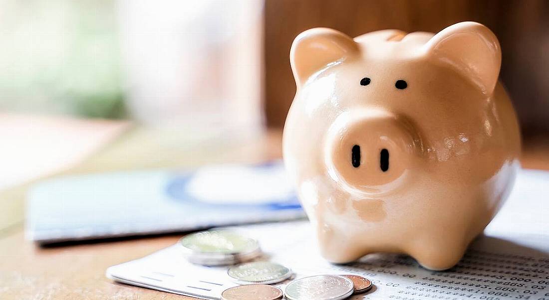 A piggy bank sits on a stack of papers with numbers on it as well as a collection of coins.