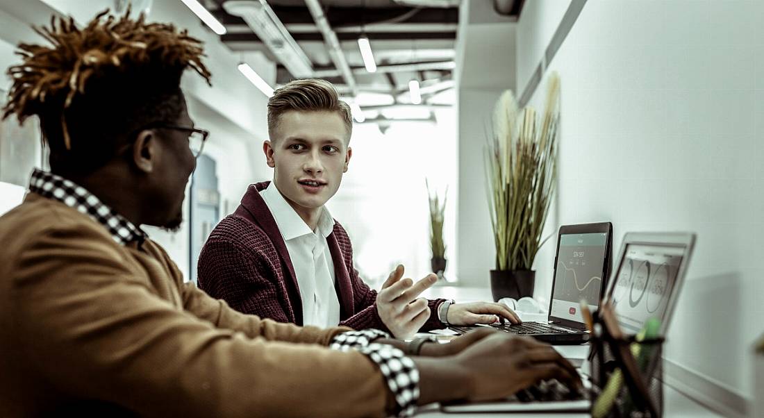 Two men working in a co-working hub standing talking while working on laptops.