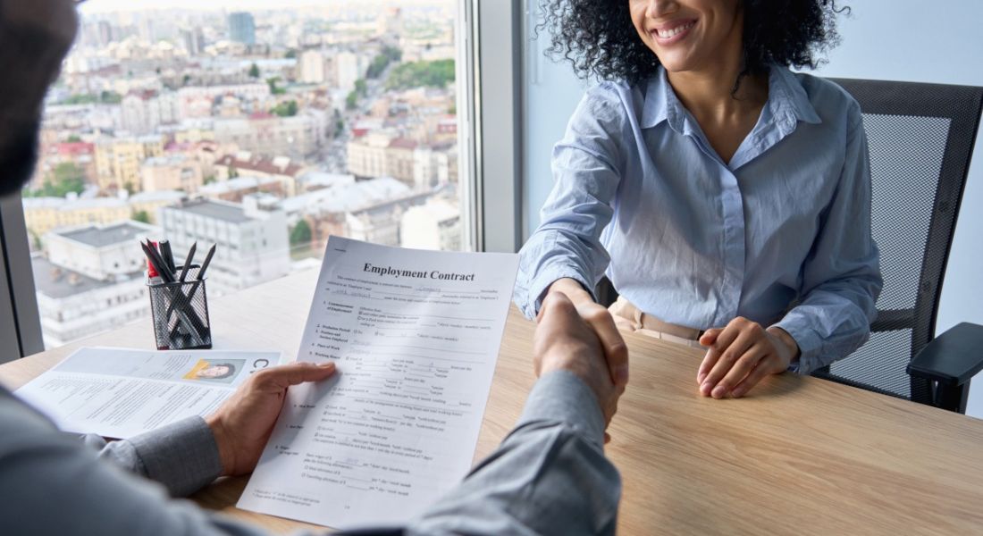 Two people shaking hands across a desk with one holding a CV.