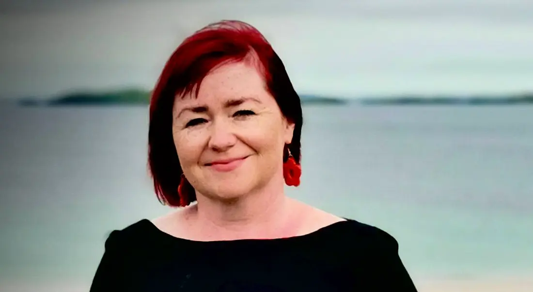 A woman with short hair smiles at the camera against the backdrop of the ocean.