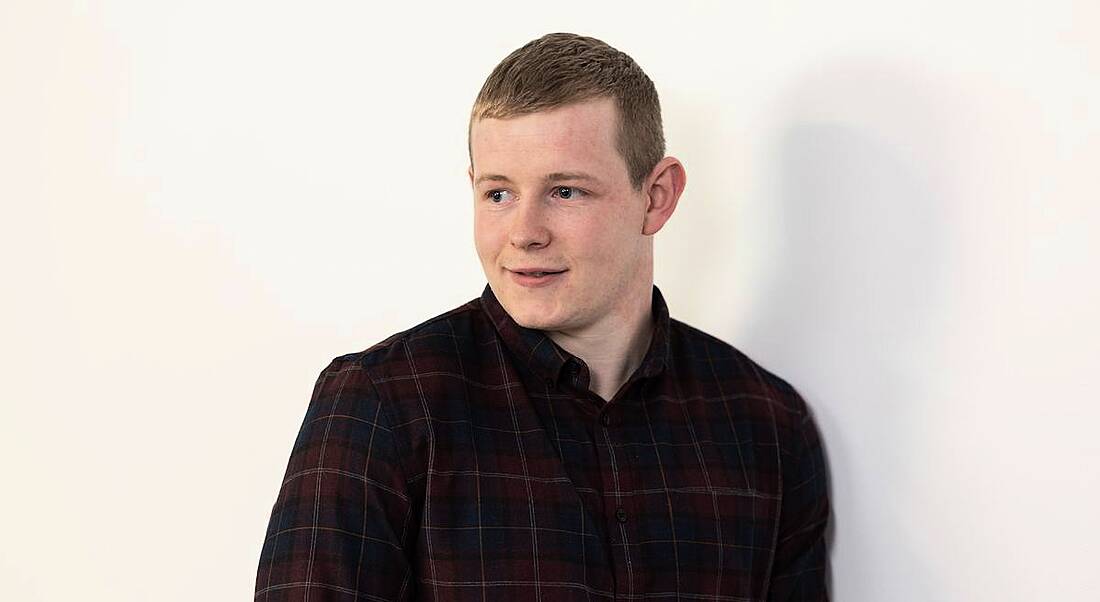 A young man in a check shirt looks off camera against a white background. He is a biopharma graduate working in BMS.