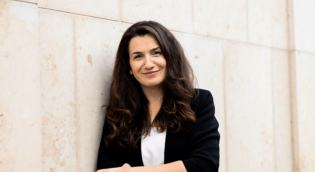 A woman with brown hair wearing a black blazer leans against a bright wall.