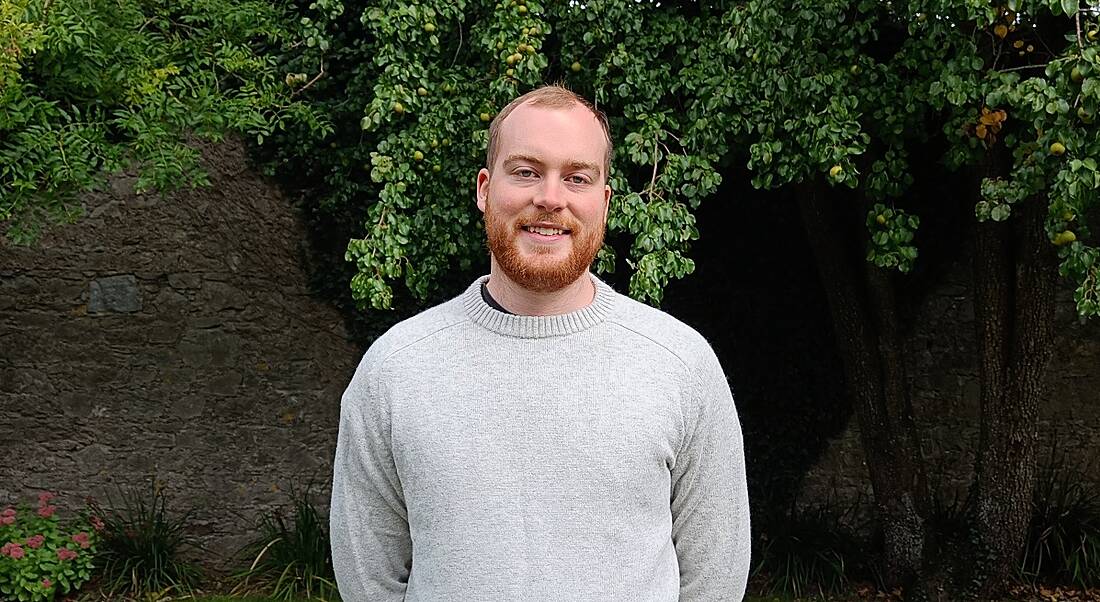 A young man in a light grey jumper smiles at the camera while standing outside.