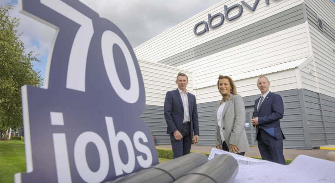 Three people stand outside the AbbVie site in Cork, beside a sign that says '70 jobs'.