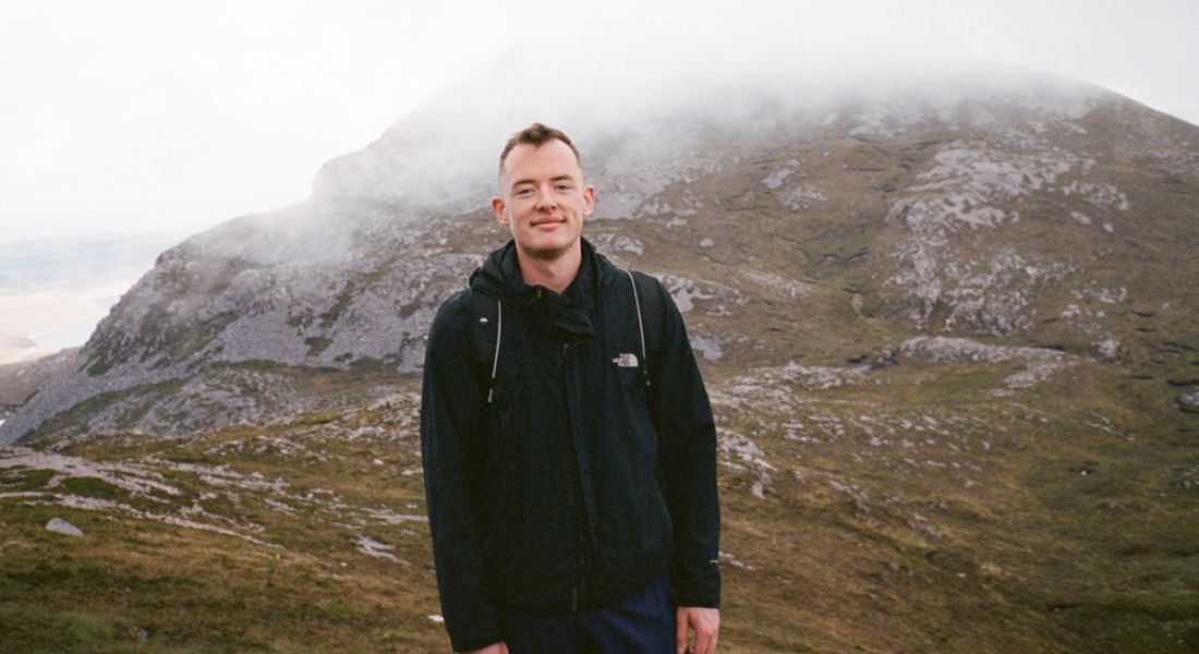 A young man stands outdoors in front of a misty mountain. He is smiling at the camera.