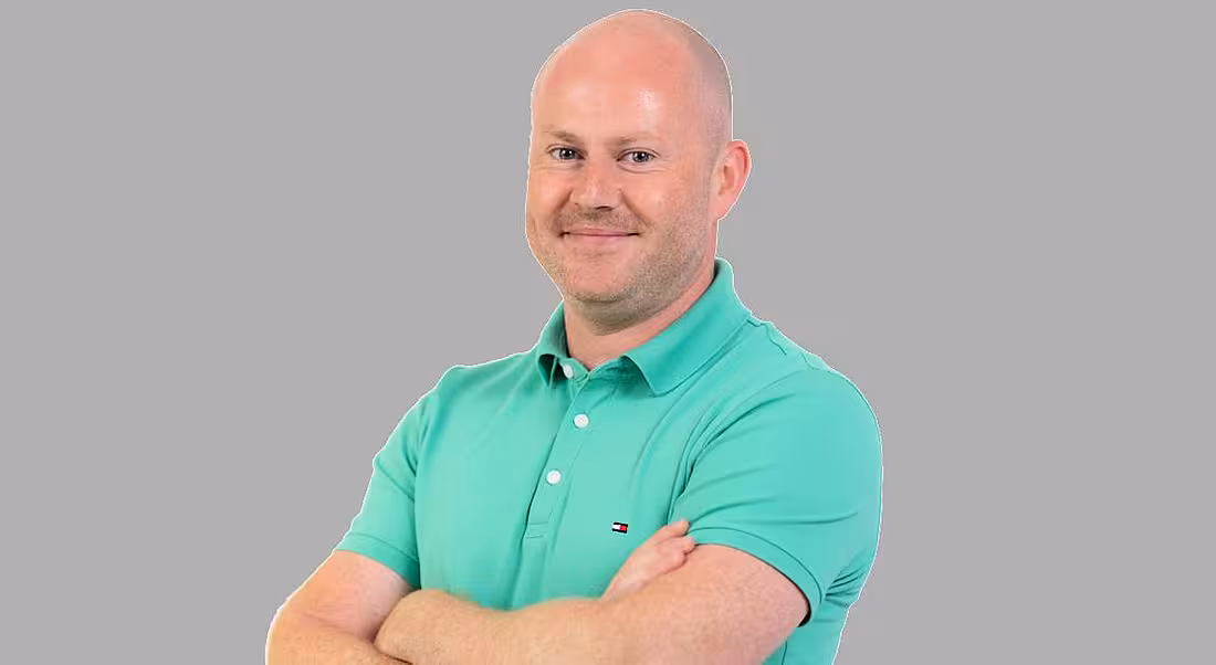 A headshot of Simon Cameron, smiling and standing with his arms folded.