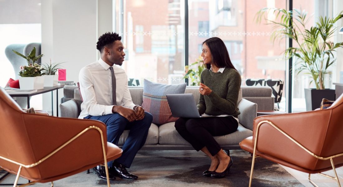 A young man and a young woman sit on a couch in a bright office having an informal stay interview.