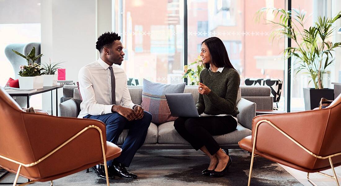 A young man and a young woman sit on a couch in a bright office having an informal stay interview.