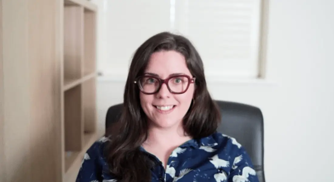 A woman wearing a blue shirt and glasses smiles at the camera during a remote video interview.