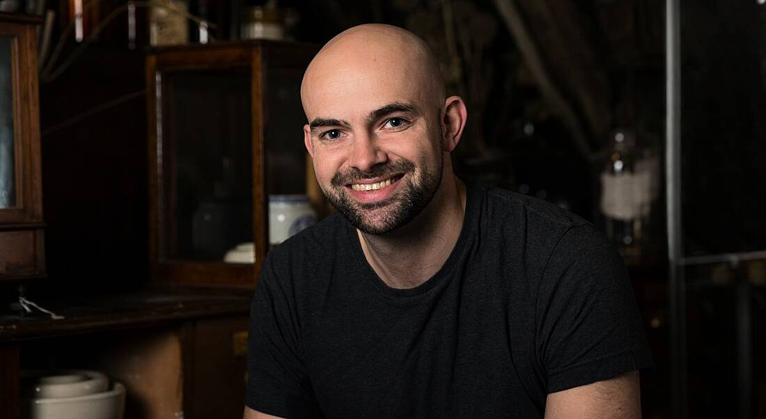 A young man wearing a grey t-shirt smiles at the camera.