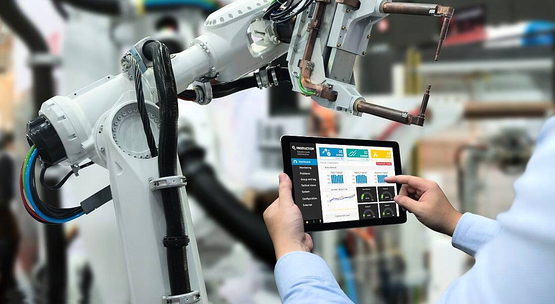 An engineer using a tablet to control a robotic arm involved in automation work, inside a factory.