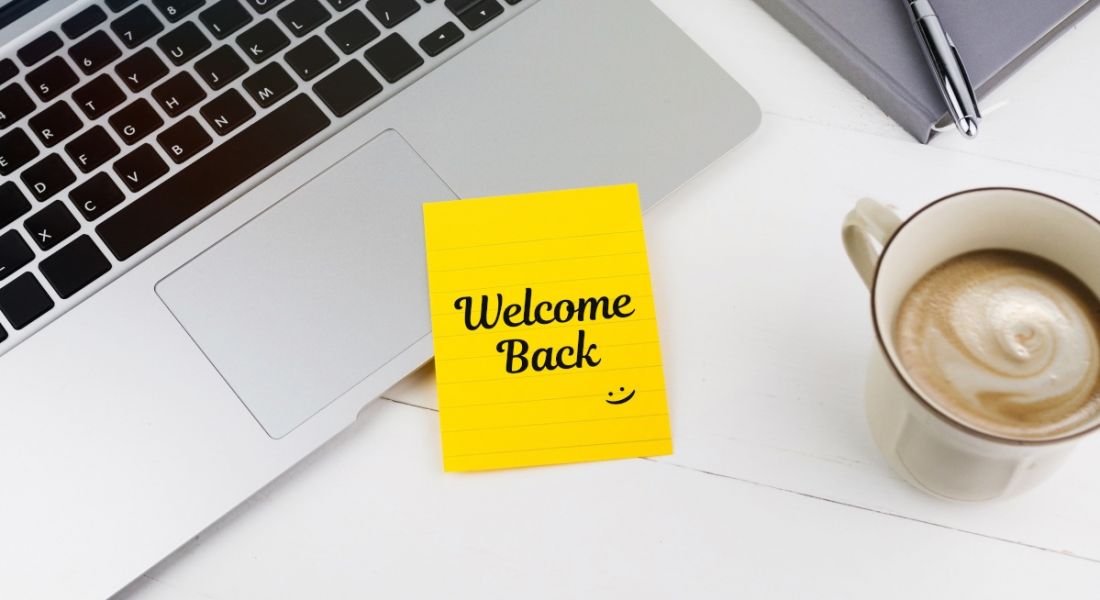 A yellow post-it note is welcoming a person back to work, sitting beside a laptop and a cup of coffee.