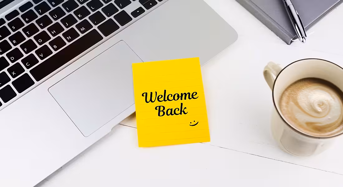 A yellow post-it note is welcoming a person back to work, sitting beside a laptop and a cup of coffee.