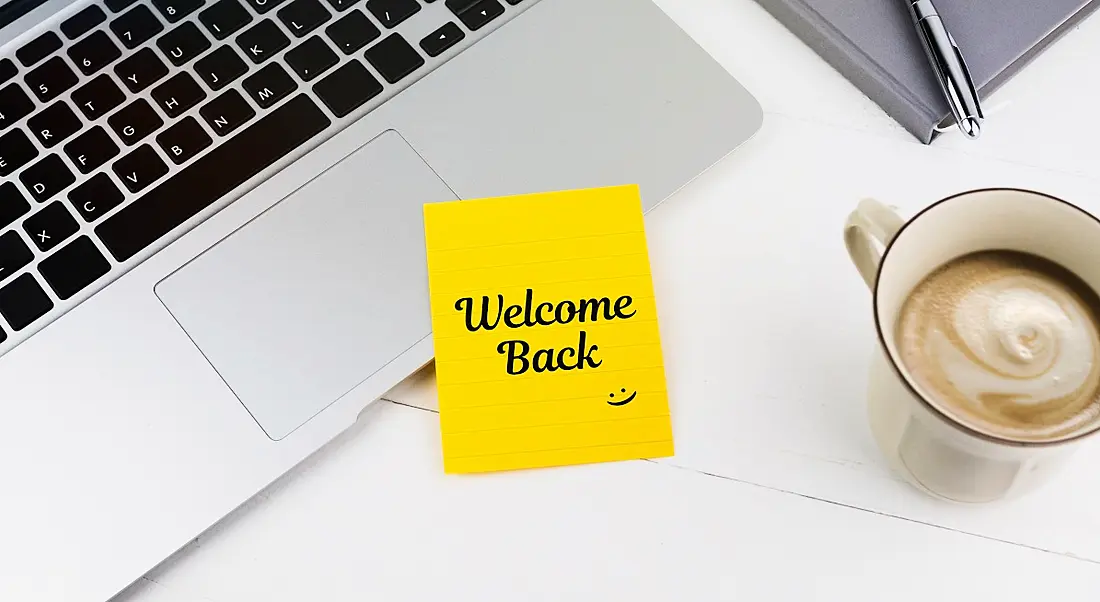 A yellow post-it note is welcoming a person back to work, sitting beside a laptop and a cup of coffee.