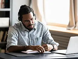 A man with glasses wearing a casual, open shirt over a t-shirt works at a computer in an office. Two co-workers are chatting in the background.