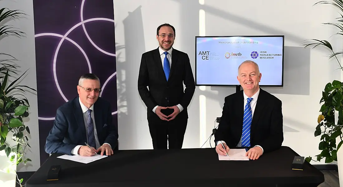 Three men, two seated at a table signing a deal with one watching them.