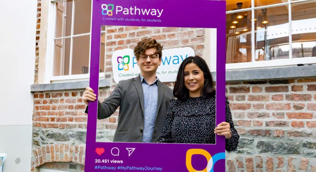 A young man and woman stand holding a large Instagram post template cut out for Pathway outside a red brick building.