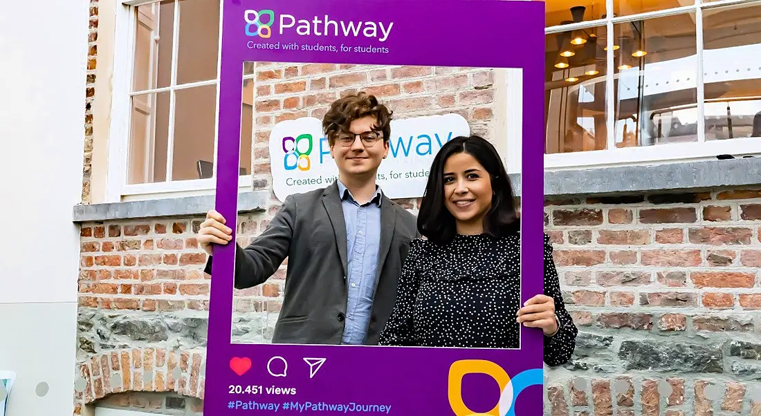 A young man and woman stand holding a large Instagram post template cut out for Pathway outside a red brick building.